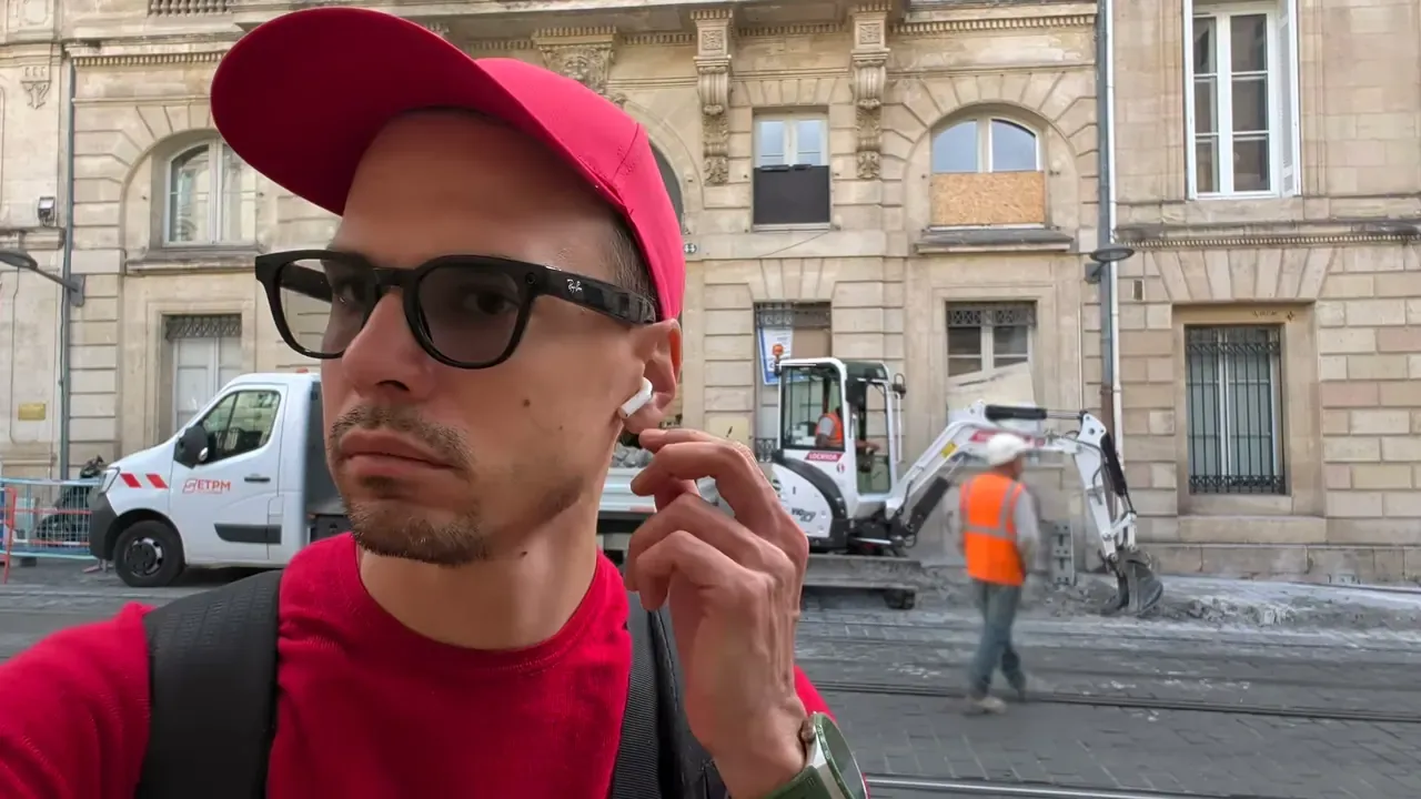 Kévin pince la tige d’un écouteur AirPods 5, devant un chantier à Bordeaux